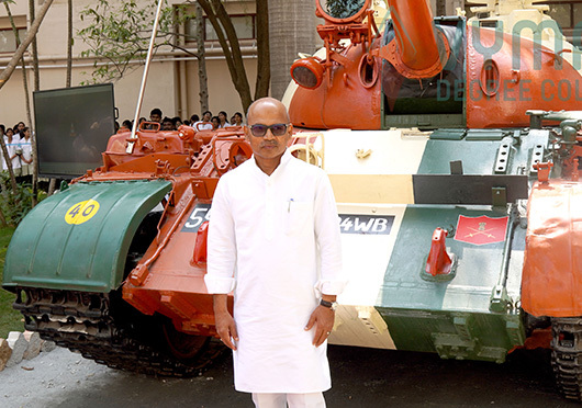 A distinguished man in white traditional attire stands in front of a decorated Indian army tank at Vymak Degree College, Bangalore.